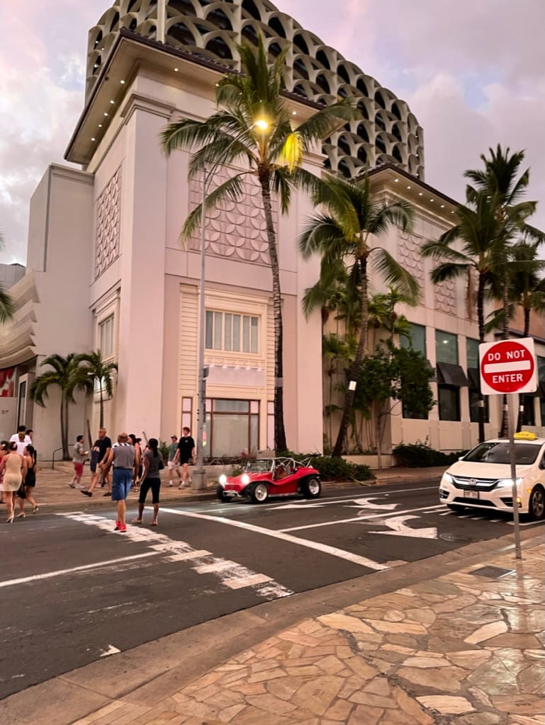 Waikiki street at dusk with palm trees, a red dune buggy, and pedestrians