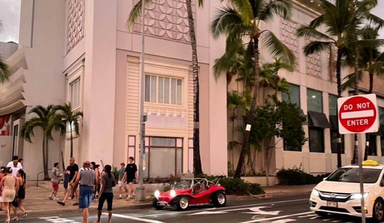Waikiki street at dusk with palm trees, a red dune buggy, and pedestrians